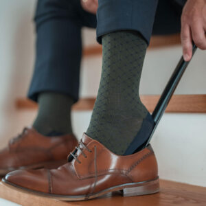 Mann zieht sich elegante braune Schuhe mit Schuhlöffel an und trägt dunkelgrüne Kompressionsstrümpfe mit Muster von xunt